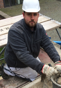 Junger Mann mit Helm kniet auf Gerüst und arbeitet konzentriert mit Mörtel.